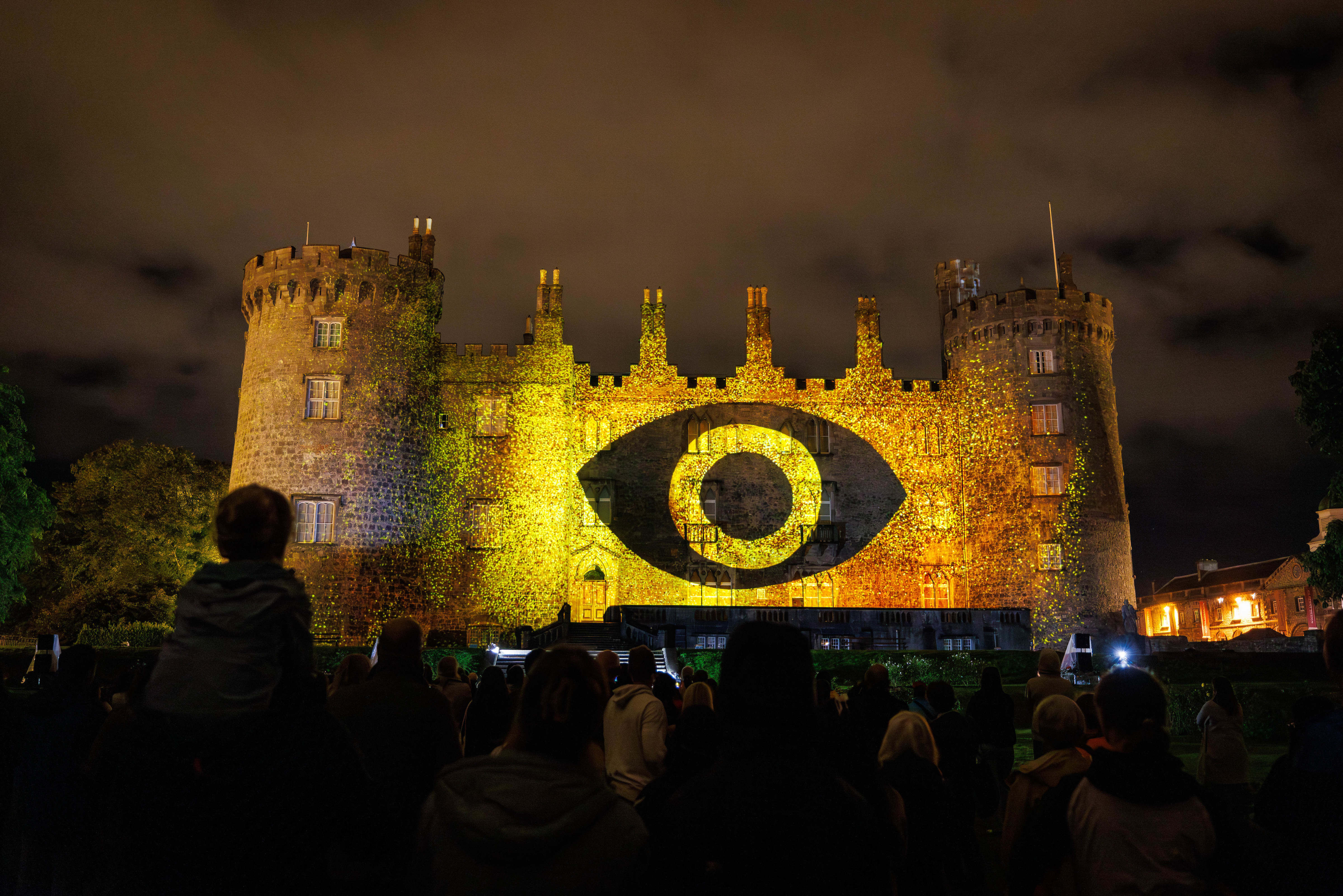 A crowd watches graphic art being projected onto Kilkenny Castle in the night.
